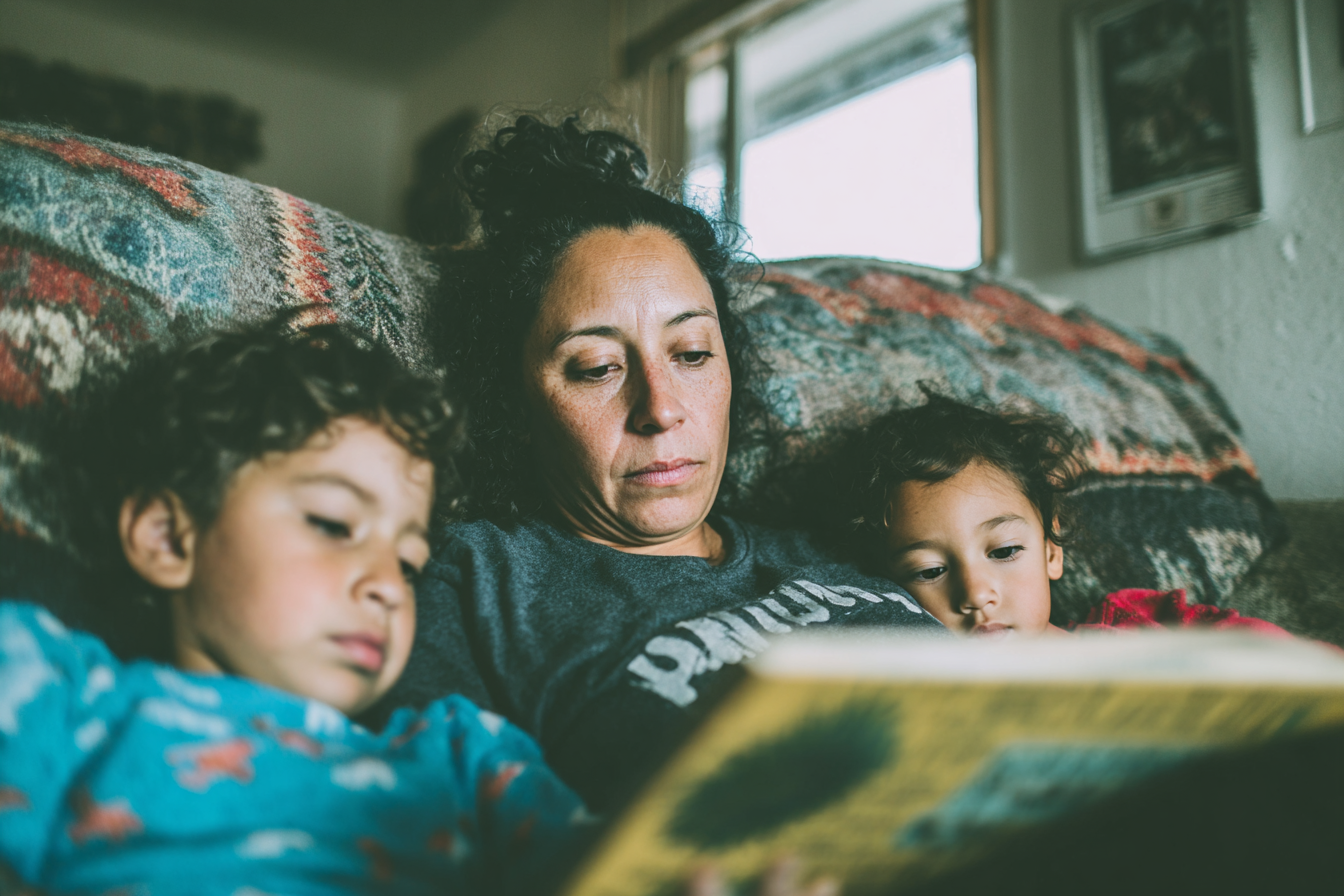 A mother reads a storybook to two children curled up beside her on a couch.