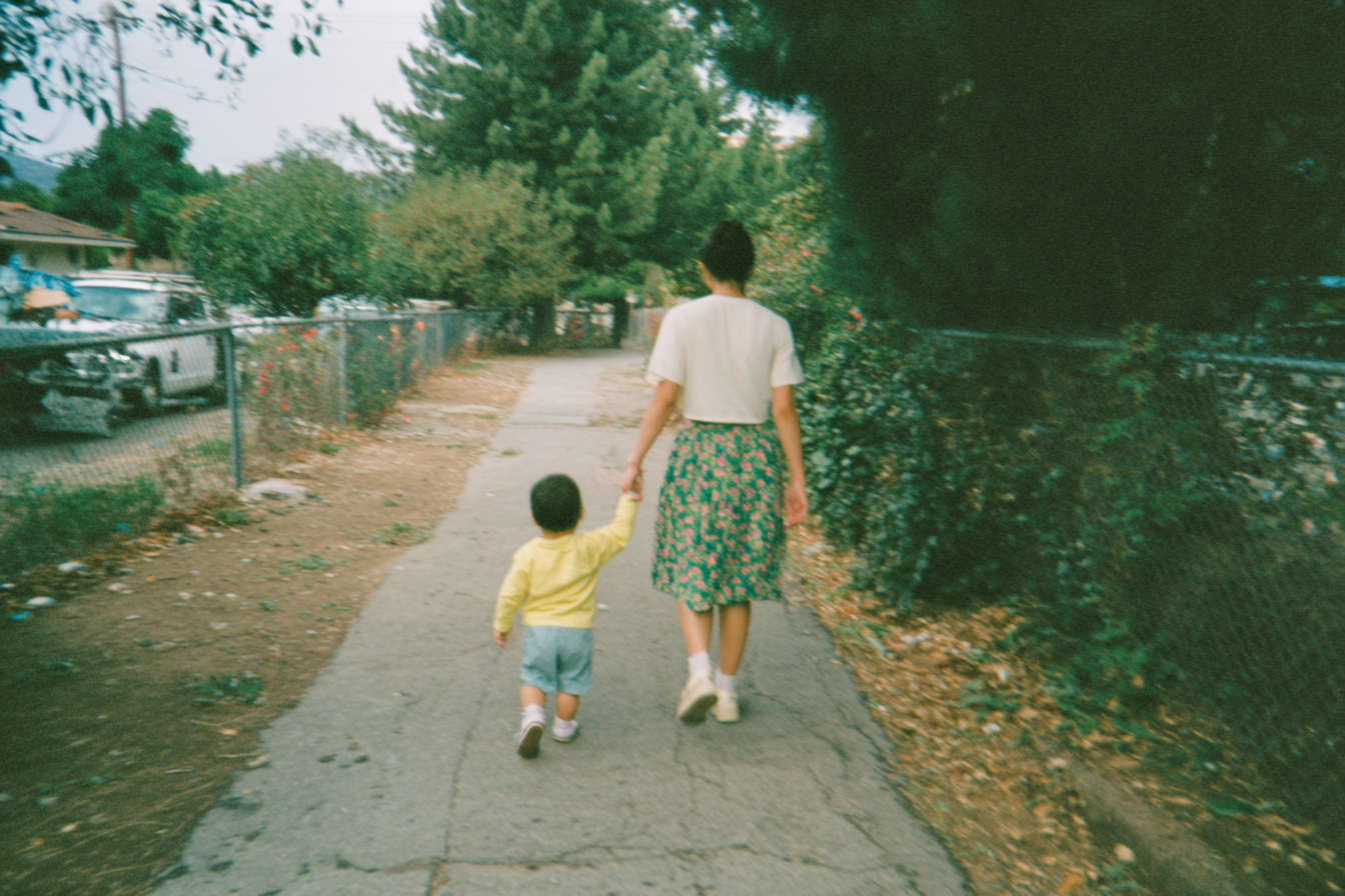 A woman walks hand-in-hand with a small child down a sunlit sidewalk.